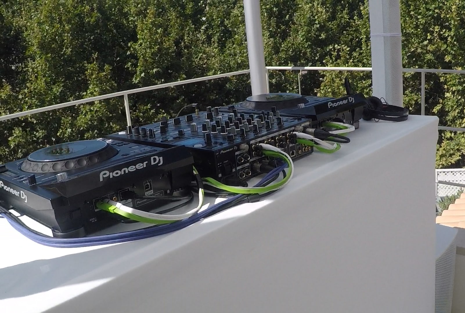 DJ equipment setup with Pioneer DJ mixers and decks on a white platform, surrounded by green cables, against a backdrop of trees.
