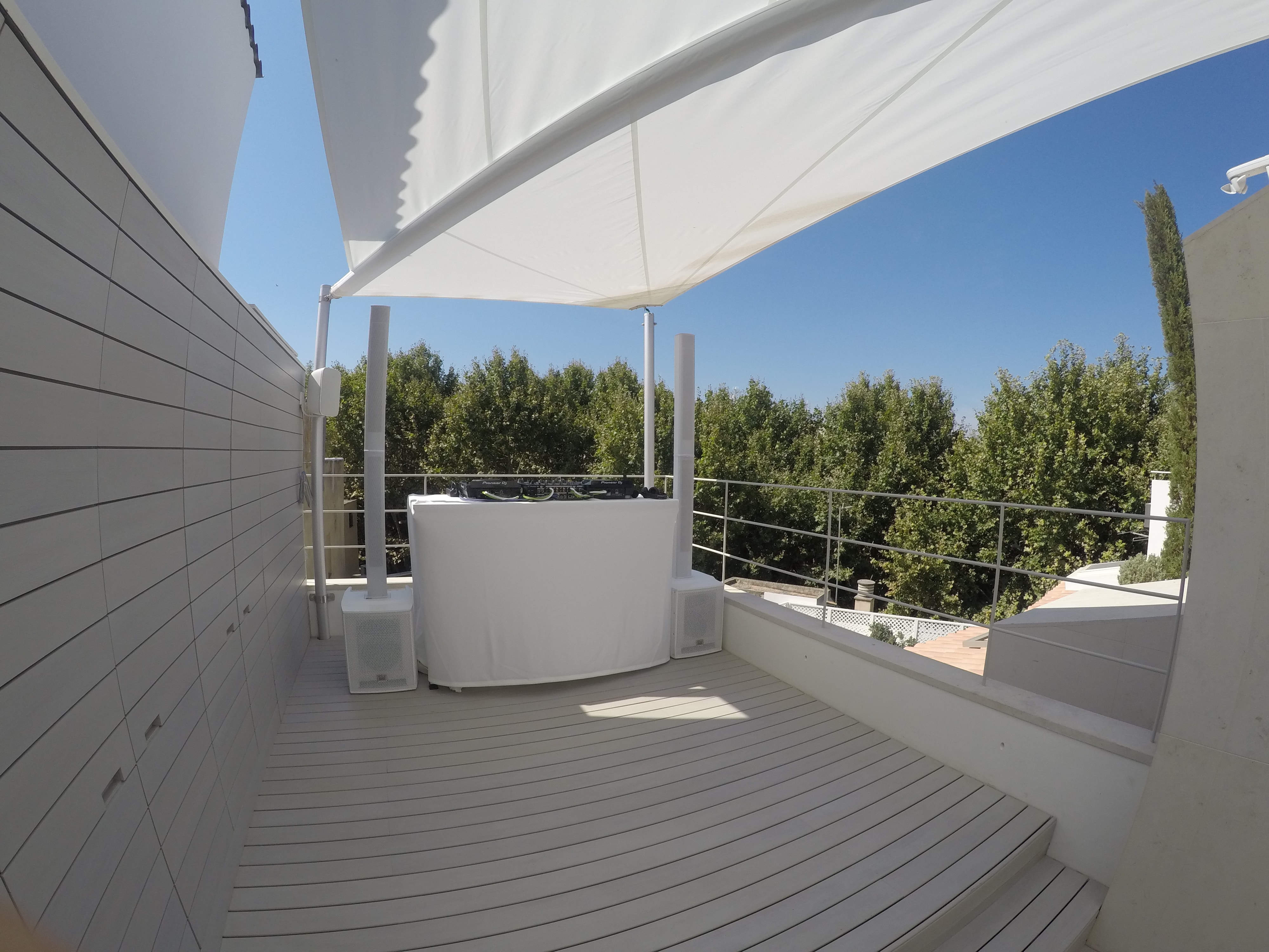 Rooftop patio with a large white canopy, a covered DJ setup, and scenic tree views in the background under a clear blue sky.