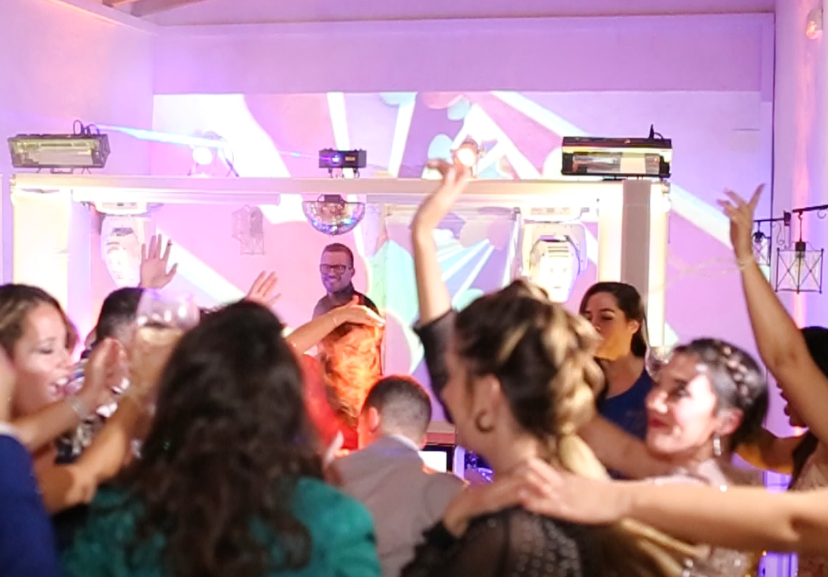 People are dancing in a lively party scene with a DJ in the background under colorful lights.