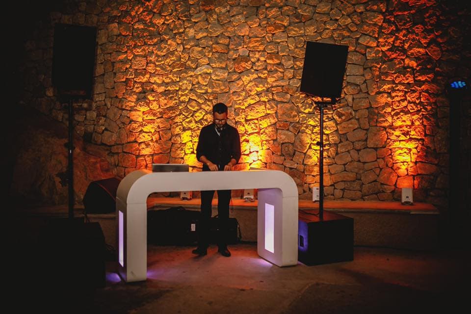 A DJ stands at a lit-up booth, performing in front of a stone wall with warm orange lighting. Two large speakers are positioned on either side.