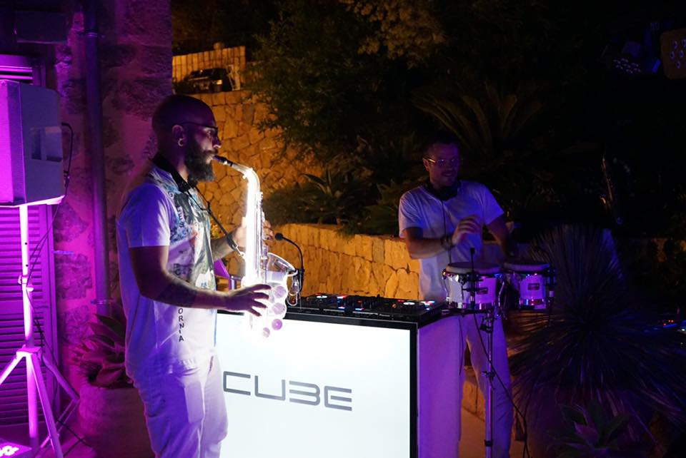 Two musicians perform at night; one plays a glowing saxophone, and the other operates DJ equipment behind a cube-shaped stand. They are outdoors, illuminated by purple lighting.