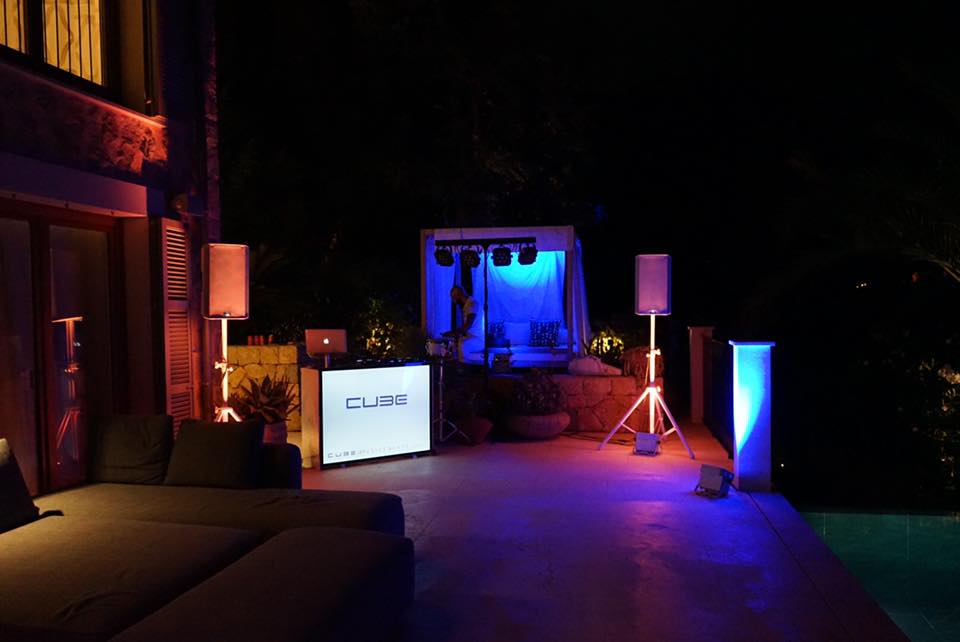 Outdoor patio setup with DJ booth, speakers, and vibrant lighting near a pool at night.