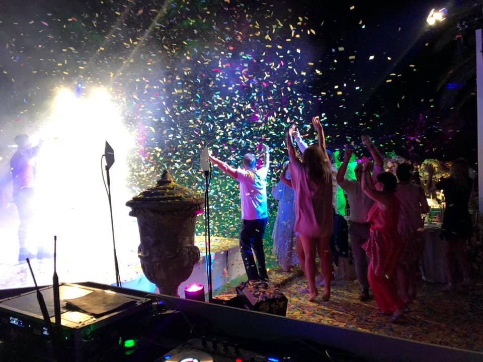 People dancing under colorful confetti at a lively party with bright lights and music equipment visible.