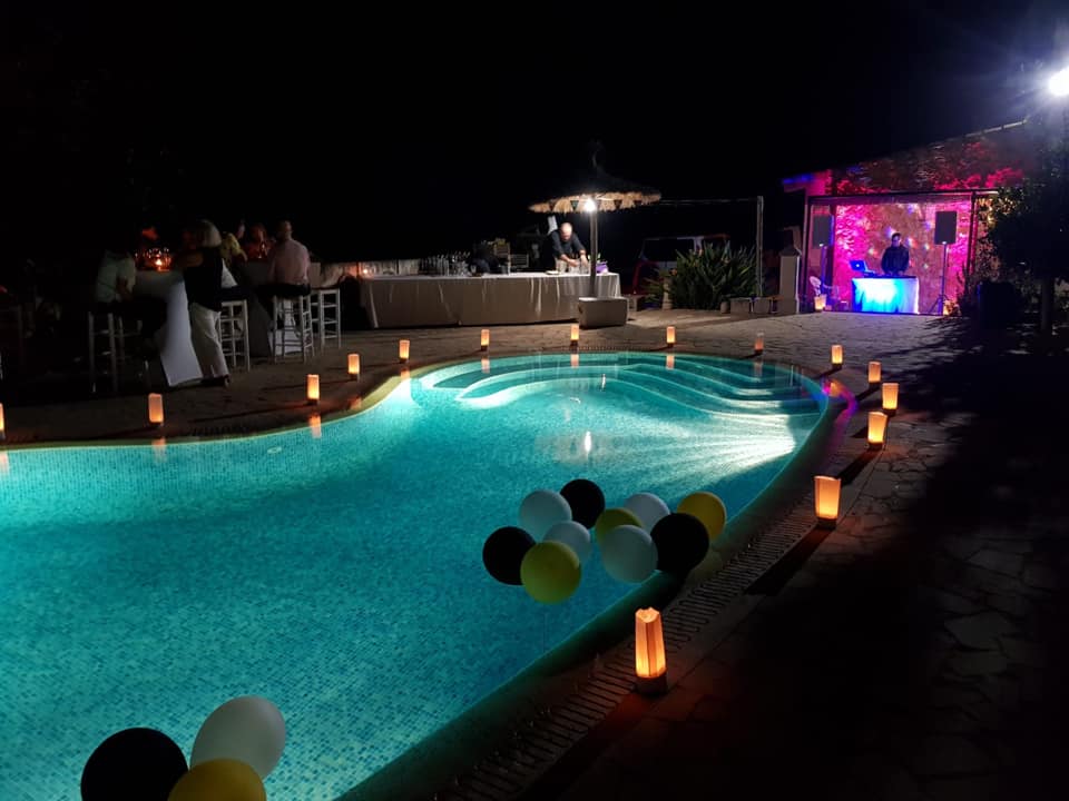 A nighttime poolside event with floating balloons and illuminated candles lining the edge. People are seated at tables in the background near a DJ booth with colorful lights.