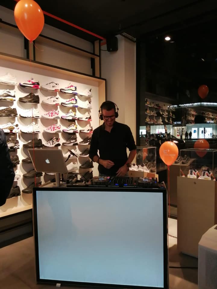 A DJ wearing headphones performs at a mixing station in a shoe store, with sneakers displayed on the wall and orange balloons visible in the background.