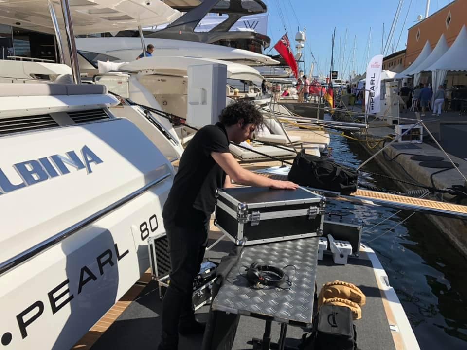 A person sets up audio equipment on a boat docked at a marina. Several other yachts and tents are visible in the background.