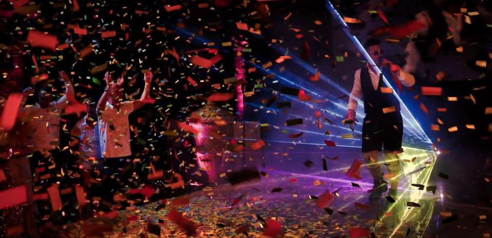 A person dances on stage surrounded by colorful confetti and laser lights, while a crowd cheers in the background.