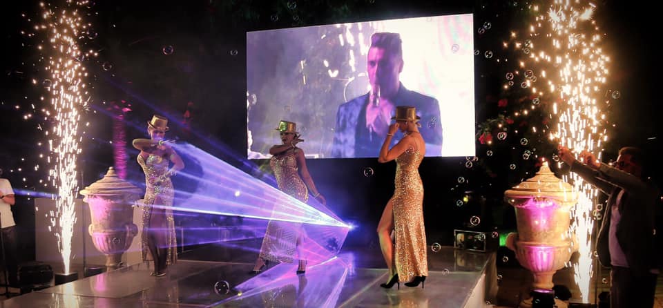 Performers in sequined outfits dance on a stage with laser lights and fireworks, in front of a large screen showing a person singing.