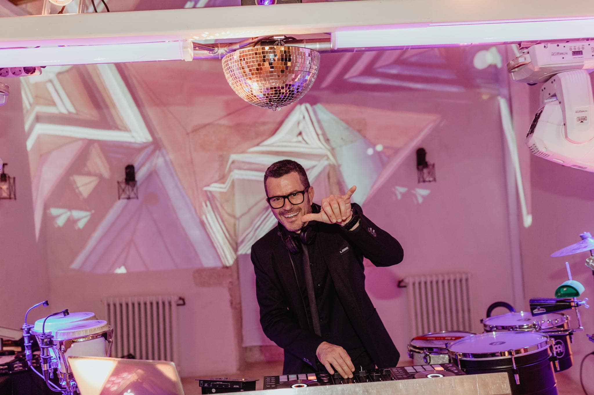 DJ smiling and playing music at a mixing station, with a disco ball and abstract projections in the background.