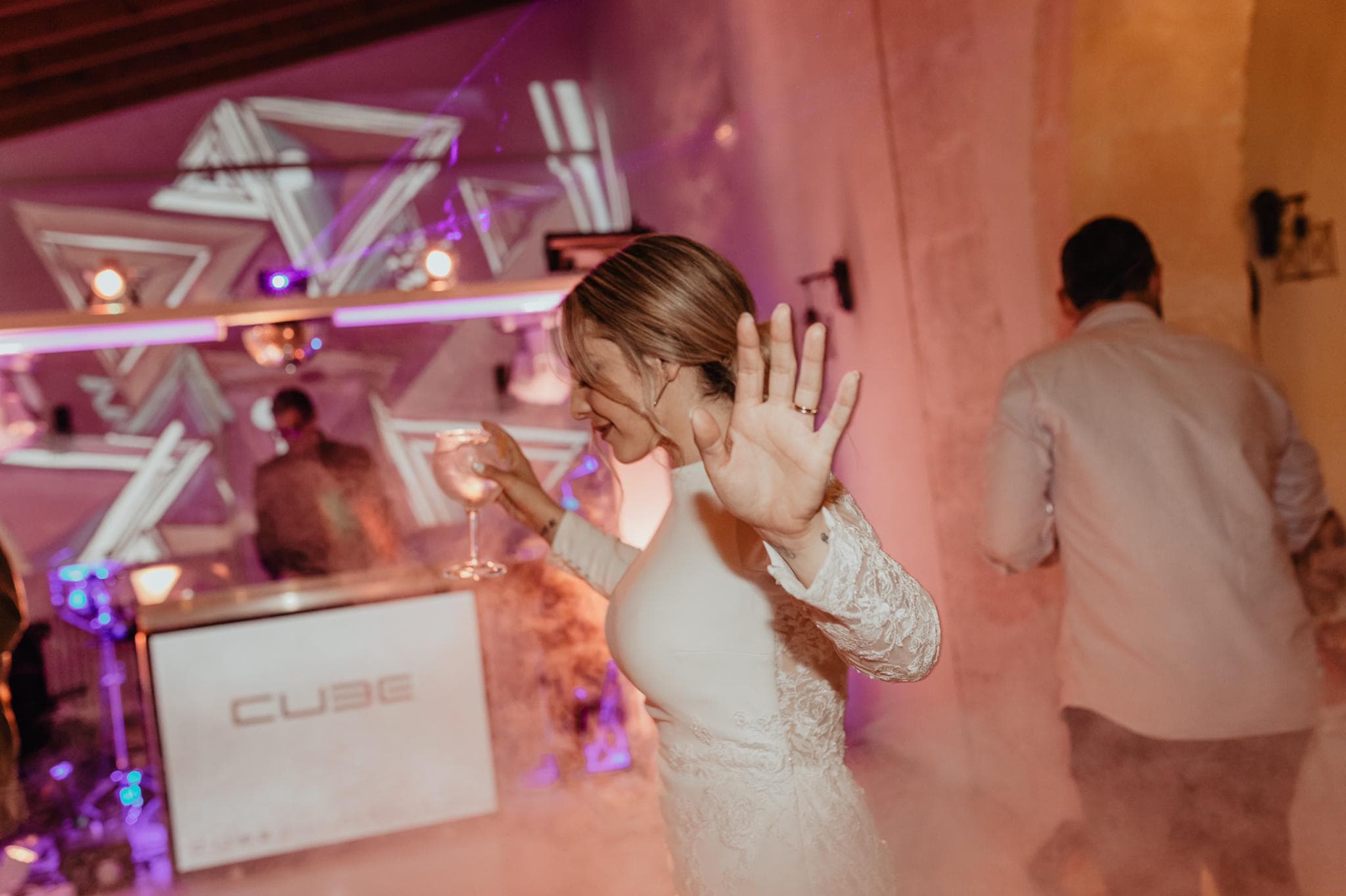 A person in a white outfit holding a drink raises their hand on a dance floor with fog and colorful lights. A DJ booth in the background features geometric light projections.