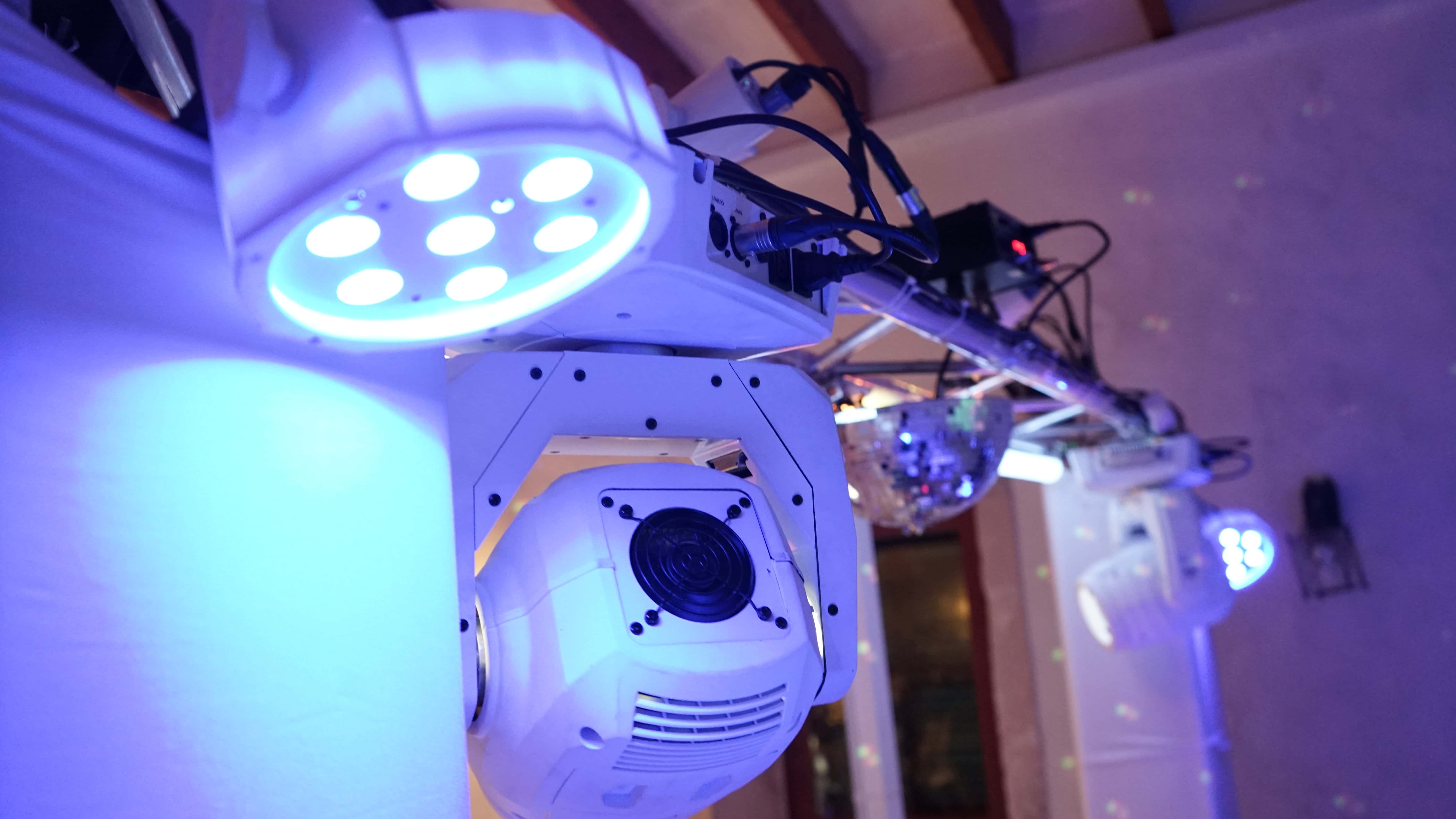 Stage lighting rig with several spotlights and equipment mounted on a metal frame, emitting blue and white light in a dimly lit space.