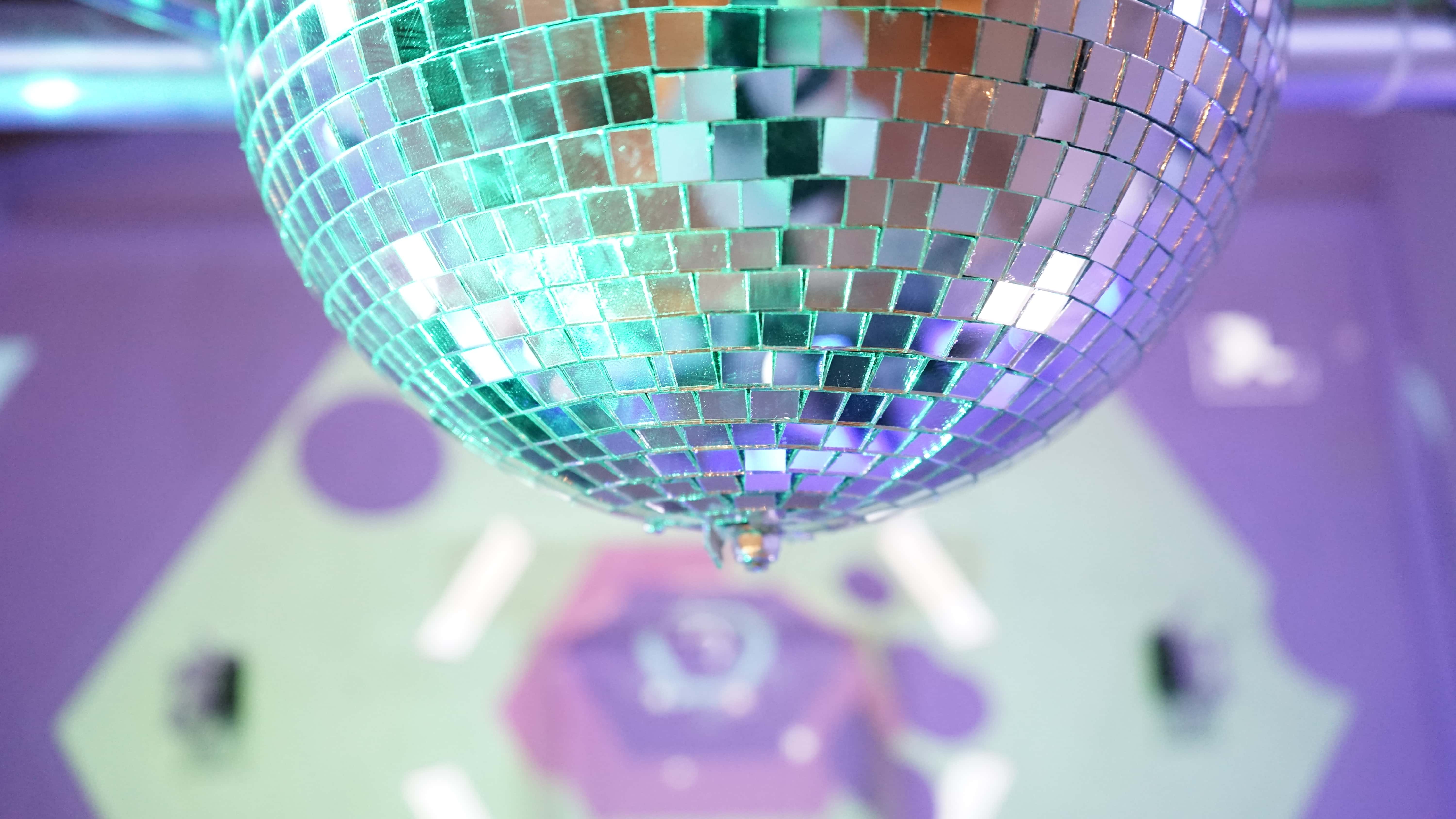 Close-up of a mirrored disco ball reflecting green and purple lights, with a blurred geometric pattern in the background.