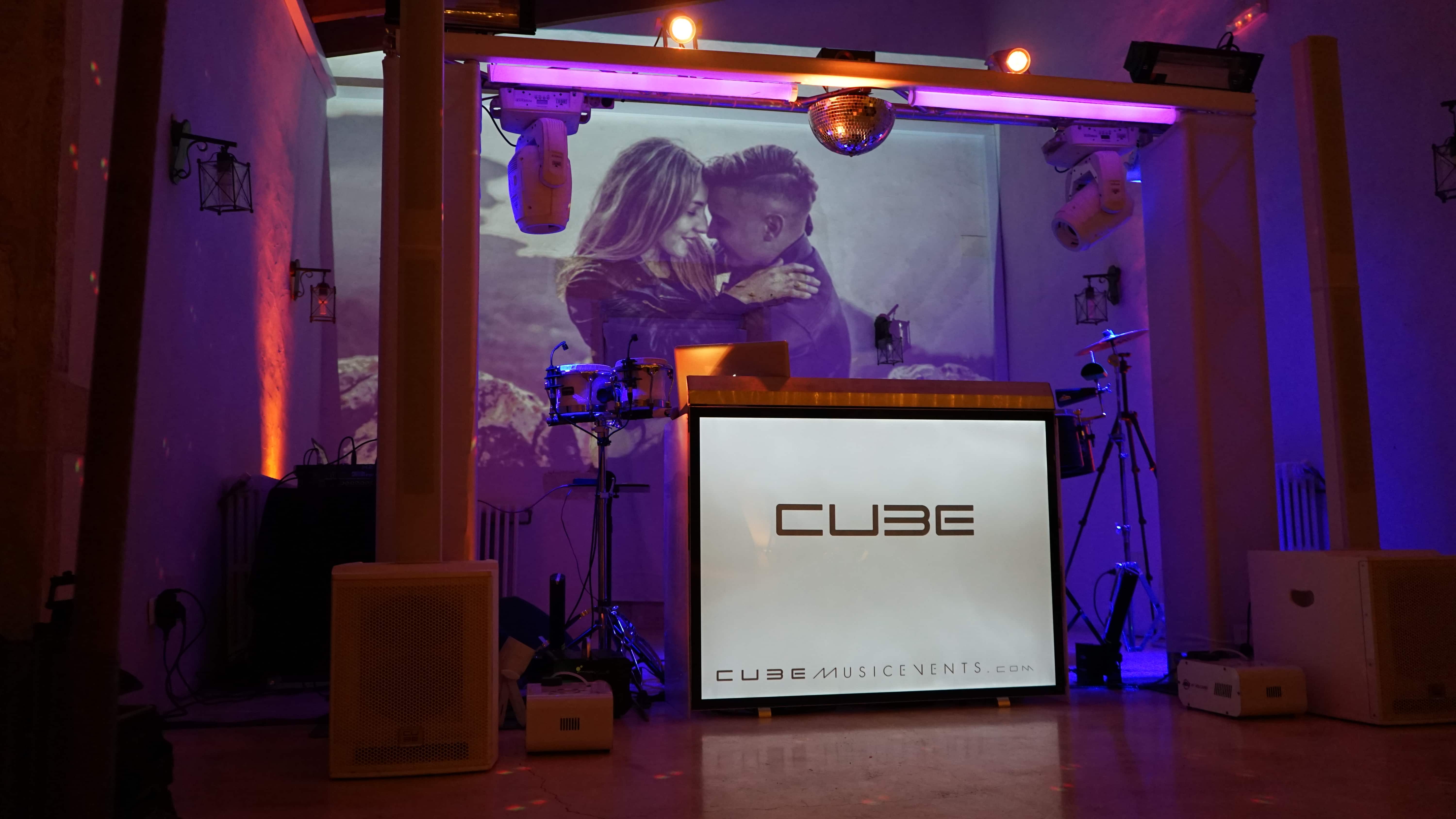DJ booth display with "CUBE" logo, featuring a large screen showing a couple embracing. Stage lights and speakers are set up around the booth.