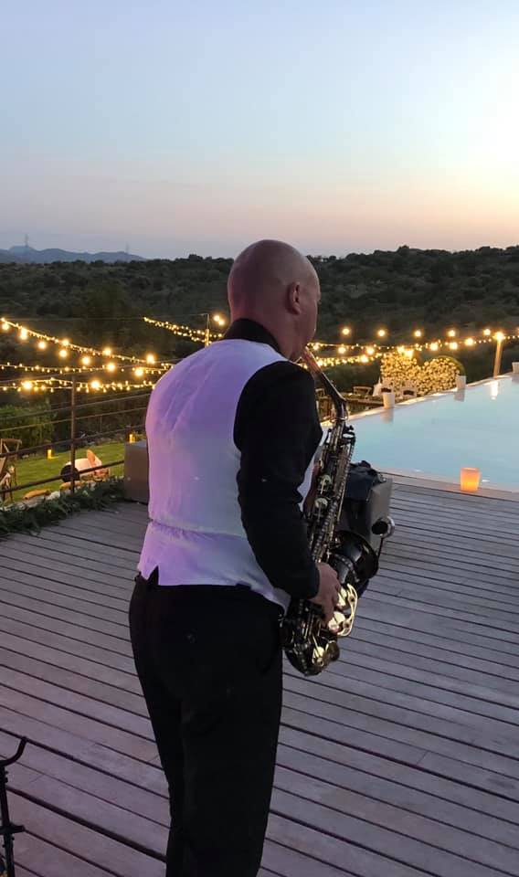 A saxophonist plays on a wooden deck near a pool at sunset, with string lights overhead.