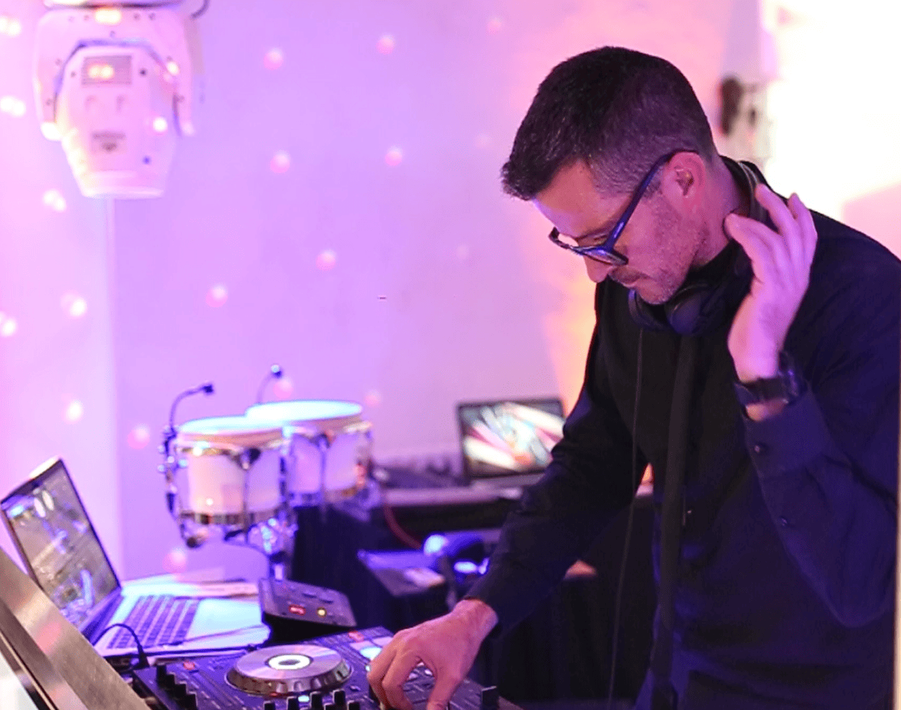 A DJ wearing headphones mixes music at a colorful, illuminated setup with laptops and percussion instruments in the background.