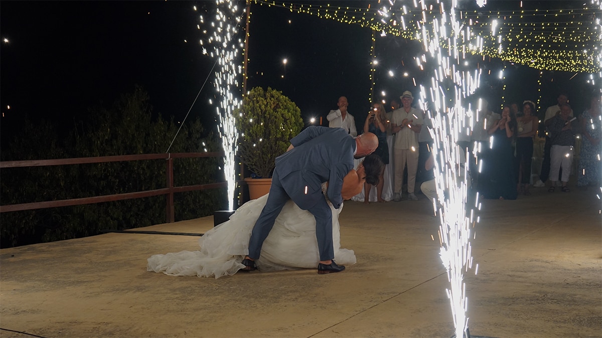 A couple dances on an outdoor patio surrounded by sparklers and lights, with guests watching in the background.