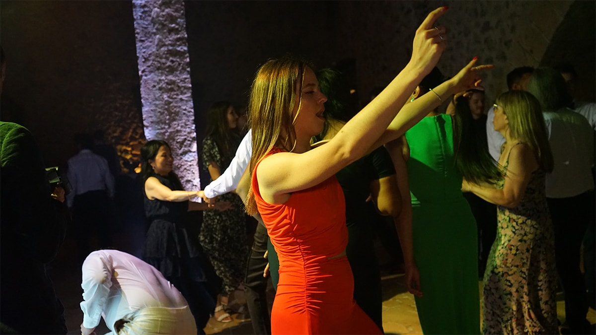 People dancing in a dimly lit room, with a woman in a red dress in the foreground raising her arms.