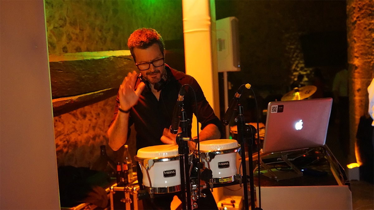 A musician playing conga drums next to a laptop in a dimly lit room.