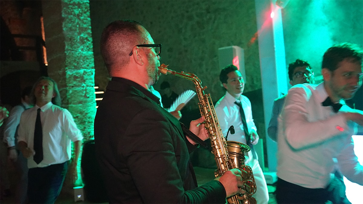 A musician in a suit plays a saxophone at a lively event, surrounded by people dancing and dressed in formal attire.