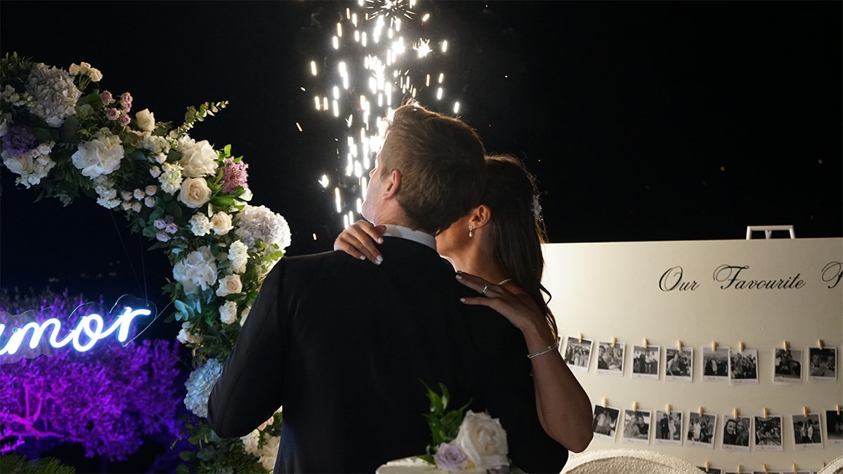 A couple embraces near a floral archway at night, with fireworks in the background and a photo display to the side.